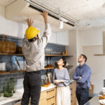 A professional HVAC technician in a yellow hard hat inspects a ceiling-mounted unit while a couple looks on, demonstrating how to choose a heating and air conditioning contractor who is thorough and communicative.