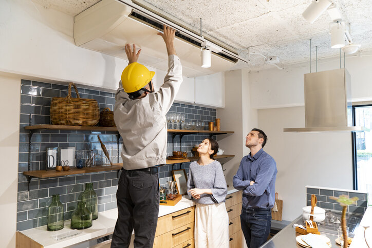 A professional HVAC technician in a yellow hard hat inspects a ceiling-mounted unit while a couple looks on, demonstrating how to choose a heating and air conditioning contractor who is thorough and communicative.